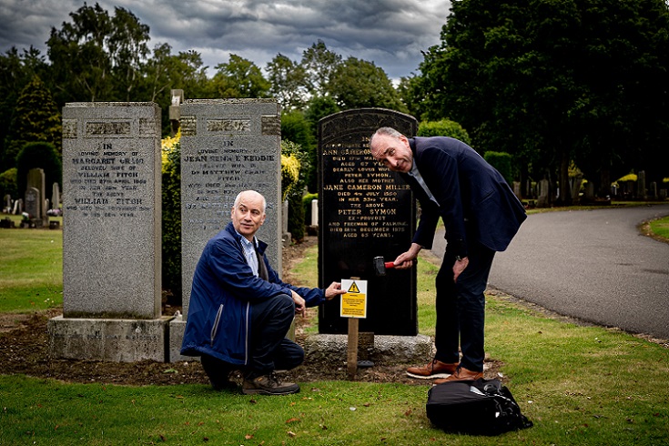 Kenneth hammers in a warning sign held by Allan at a headstone in need of repair. A photograph of the headstone is then added to PlotBox, with a further picture uploaded once repairs are complete. 